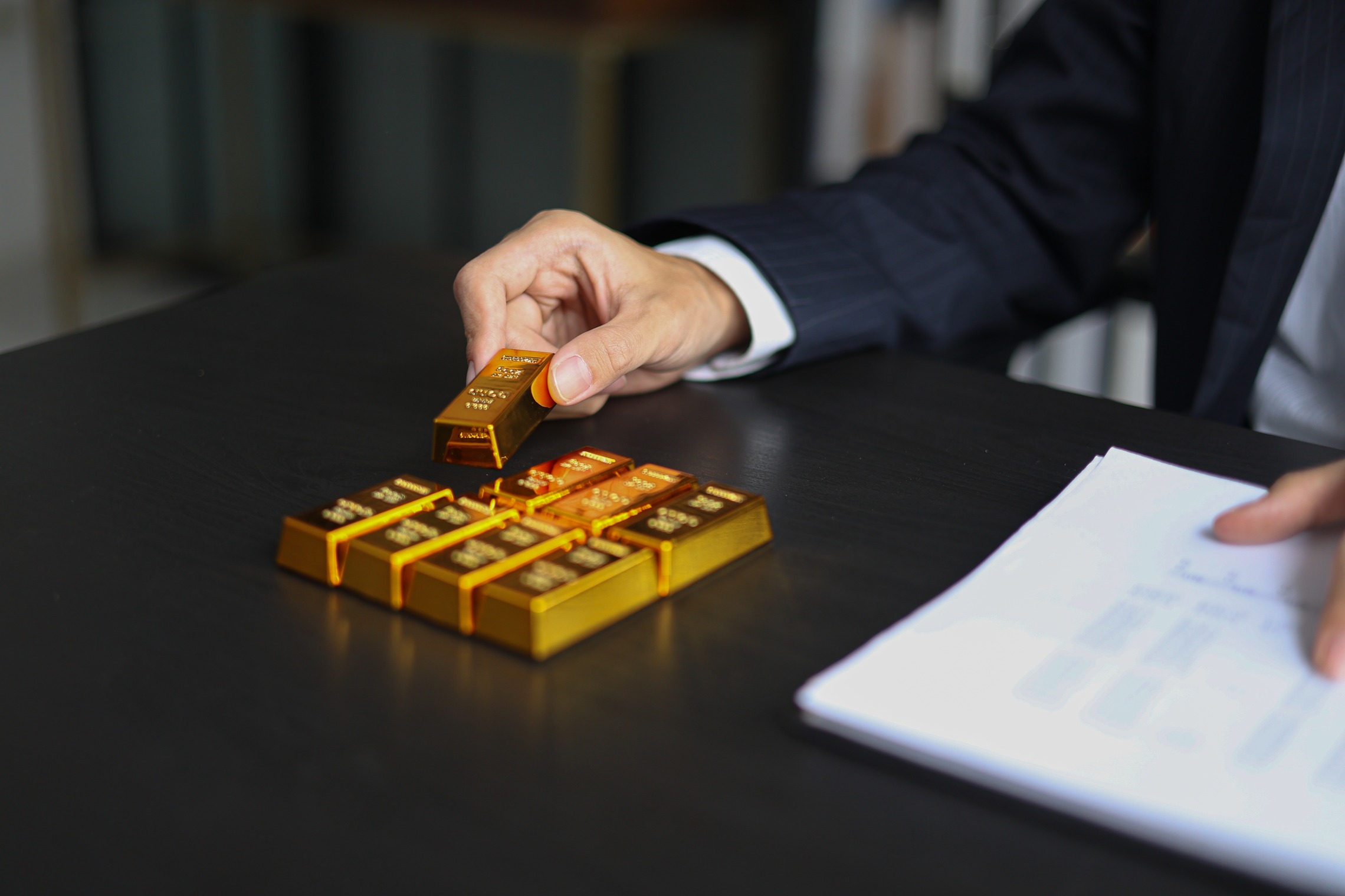 In Edelmetalle investieren - einfach bestellt, sicher gekauft Das Bild zeigt eine Hand, die auf einem Tisch Goldbarren stapelt.